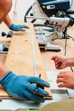 Gloved hands position measuring tape along lumber board beside miter saw in.. Stock Photos