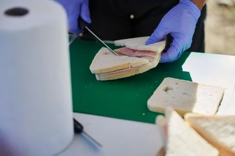 Gloved Hands Slice Ham Sandwich On Green Cutting Board Outdoors Foto stock