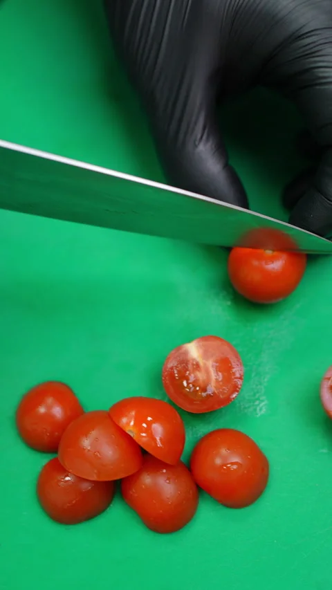 Gloved hands slicing cherry tomatoes on a green cutting board, then adding them Video stock 309119621