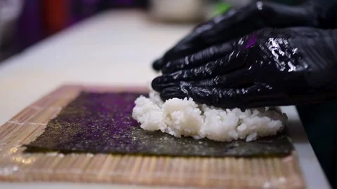 Gloved hands spreading sushi rice on a nori sheet placed on a bamboo mat Stock Footage 293491633