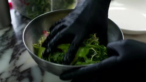 Gloved Hands Tossing Fresh Salad Greens in Metal Bowl on Marble Kitchen Counter Stock Footage 337652772
