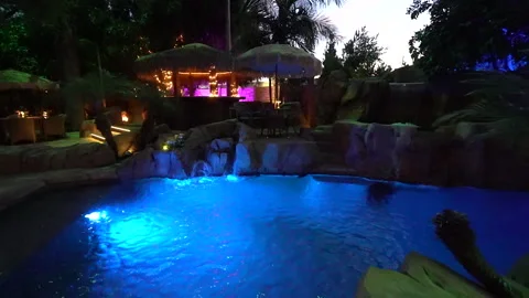 Glowing blue pool with waterfall and thatched tiki bar at night Stock Footage 345540074