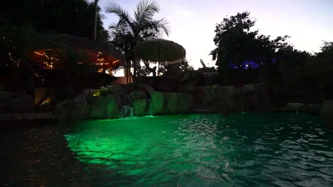 Glowing green pool with rock waterfall and tiki hut at dusk Stock Footage 345536793