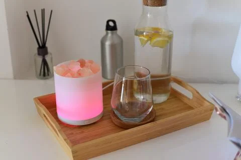 Glowing salt lamp diffuser, drinking glass, lemon water bottle on wooden tray Foto stock