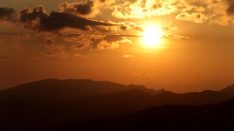 The glowing sun descends behind rugged mountains in a dramatic evening sky. Stock Footage 325593954