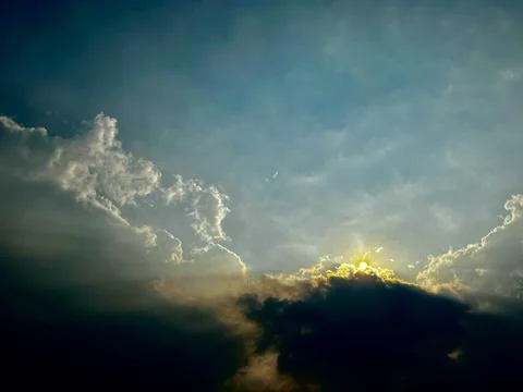 Glowing sun shining through dark clouds in dramatic blue sky during sunsite Stock Photos