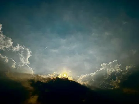 Glowing sun shining through dark clouds in dramatic blue sky during sunsite Stock Photos