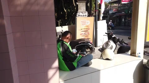 Gojek Delivery Driver Resting and Checking Smartphone on Street Bench in Ubud Stock Footage 342296771