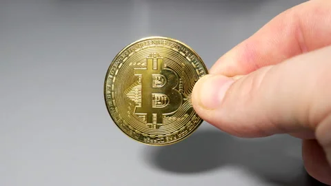 Gold bitcoin coin held between fingers in hand on gey table Stock Footage 161376317