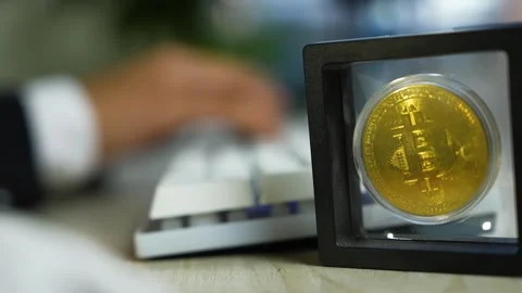 Gold bitcoin coin in protective display case foreground with blurred keyboa.. Vídeo Stock 329177002