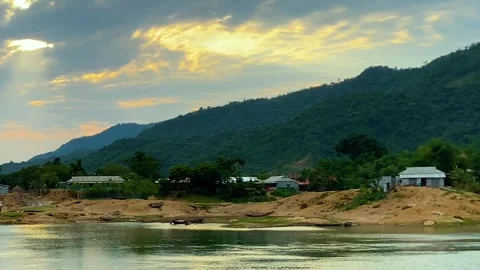 Golden light breaking through clouds over hillside village with calm lake at Vidéo 333020254