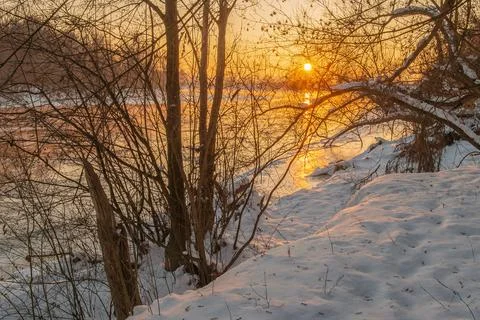 Golden rays of sunlight emerge, casting a warm glow over the frozen river Stock Photos