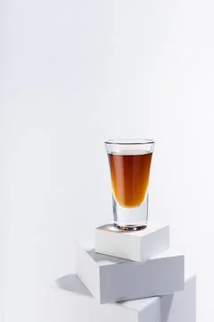Golden scotch in shot glass in sunlight with shadows on white square podium w Stock Photos