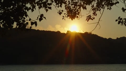 The golden setting sun bursts behind hills above a calm river Stock Footage 339630575