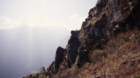 Golden sun rays stream across a rugged mountain cliff face overlooking a hazy Stock Footage 344177253