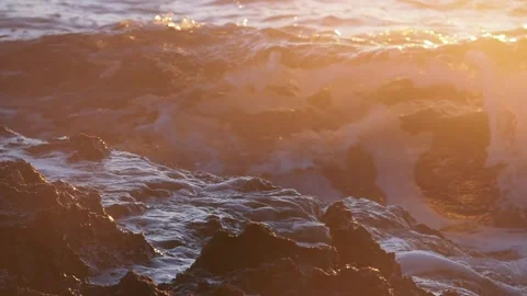 Golden sunlight illuminates ocean waves crashing against dark rocky shore Stock Footage 337414996