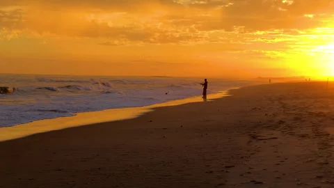 Golden sunset casts a warm glow over the beach as a fisherman stands by the wate Stock Footage 291387667