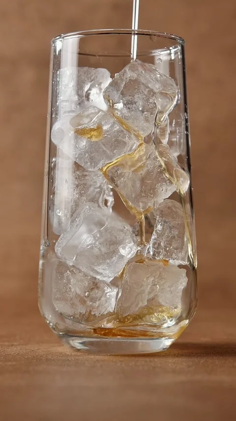 Golden syrup pours over ice cubes in tall glass, coating chilled surface during Stock Footage 343496002