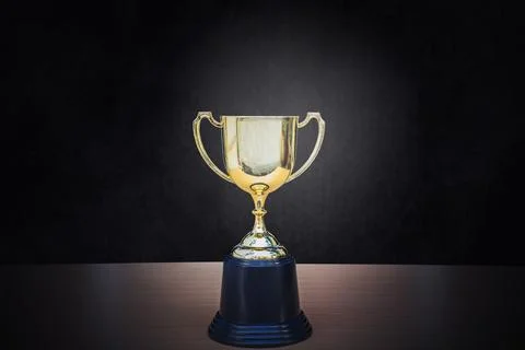 Golden trophy placed on top of old wooden table in front of dark background.. Stock Photos
