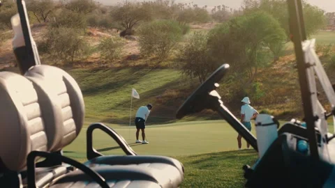 Golfer sinking a putt on a desert green framed through a golf cart in warm Stock Footage 340130126