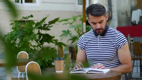 Good-looking 30-aged modern man reading interesting book in the comfortable cafe Stock Footage 112350599