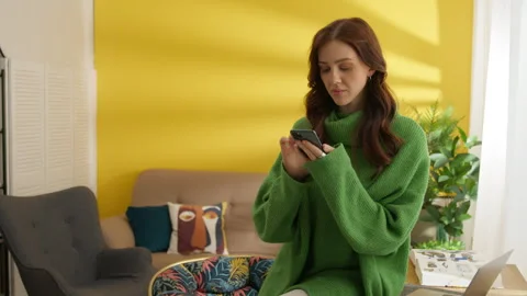 Good-looking brunette sitting on the edge of table in room, browsing cell phone Stock Footage 245465895