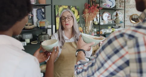 Good-looking friendly grey-haired mature seller proposing beautiful utensils to Stock Footage 143981093