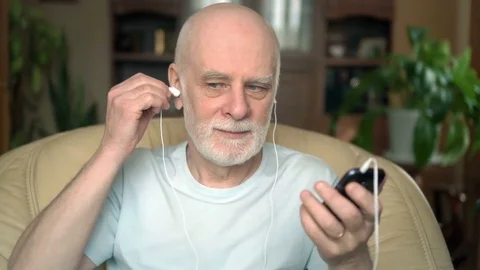 Good-looking senior man sitting in chair at home. Listening music on smartphone Stock Footage 74814964