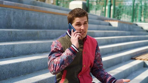 Good-looking young man talking on mobile phone on concrete stairs outside Stock Footage 128509652