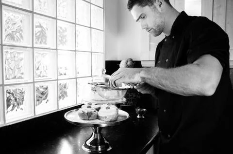 Good loooking baker busy dusting icing sugar onto iced buns Stock Photos