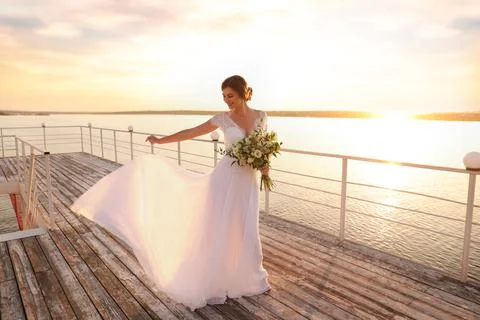 Gorgeous bride in beautiful wedding dress with bouquet near river on sunset Stock Photos