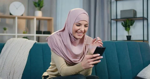 Gorgeous satisfied smiling young arabic woman in hijab sitting on the couch at Stock Footage 150110511