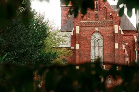 Gothic red brick cathedral facade framed by lush green foliage under soft n.. Stock Photos