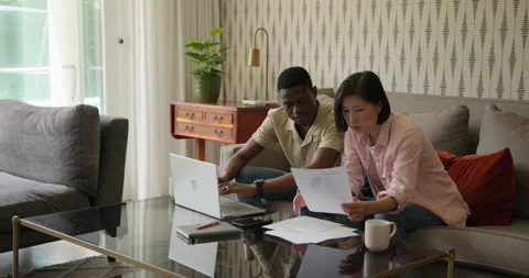Grabbing documents, Diverse couple comparing charts on laptop on sofa for Stock Footage 316614752