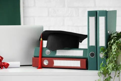 Graduation cap with red tassel on a stack of binders diploma scroll and lapto 스톡 사진