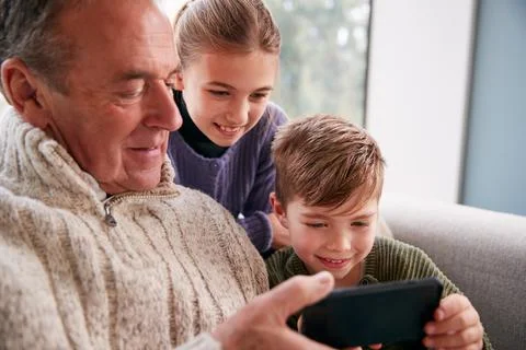 Grandchildren On Sofa At Home Showing Grandfather How To Use Mobile Phone Stock Photos