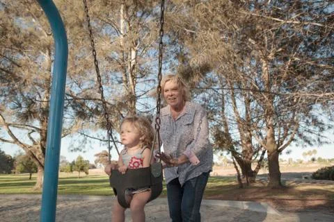 Grandmother pushing granddaughter on swing at playground Stock Photos