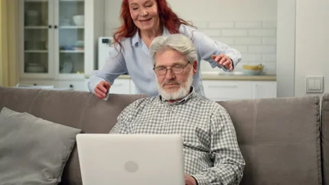 Grandpa is sitting on the couch using a laptop. Grandmother comes up to him and Stock Footage 183873743