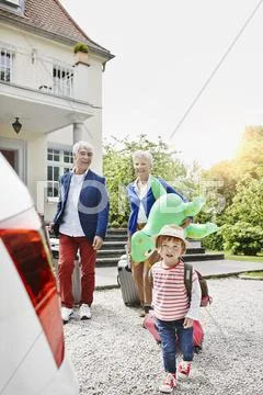 Grandparents With Granddaughter And Luggage On Driveway