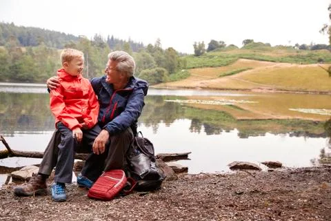 Grandson sitting on his grandfather's knee at the shore of a lake smiling, La Stock Photos