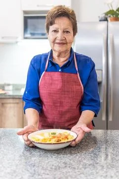 Granny looking at camera offering a soup plate in the kitchen Stock Photos