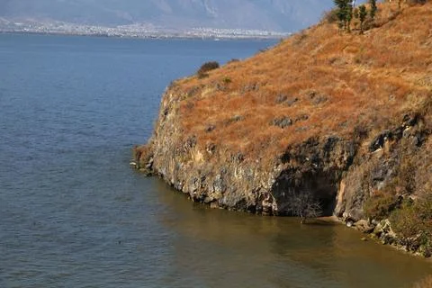 Grassy cliff sloping down to a lake with a small shore cave 스톡 사진