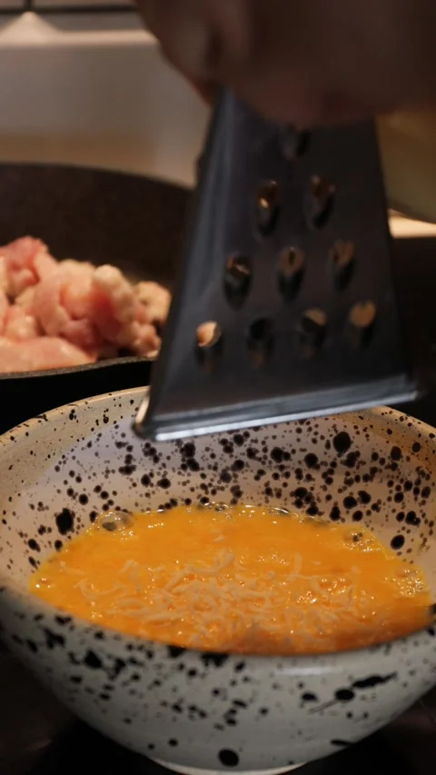 Grated parmesan falling into beaten eggs in speckled bowl with bacon frying Stock Footage 314267069