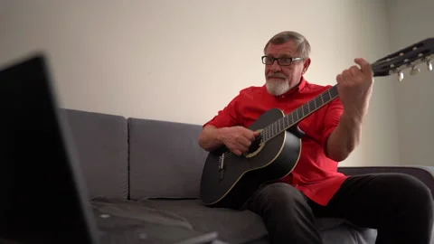 Gray-bearded pensioner with glasses playing guitar at home. Mature man learns to Stock Footage 149032314