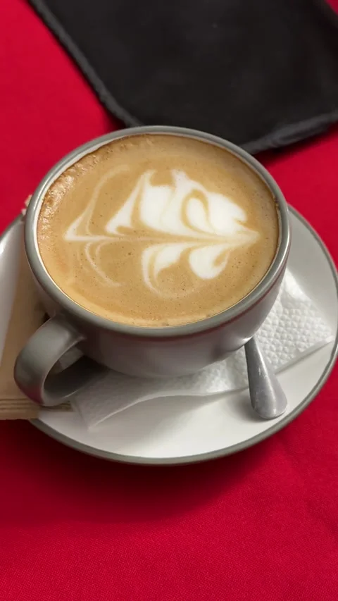 Gray ceramic mug of latte coffee with abstract foam art on red tablecloth cozy Stock Footage 345729040
