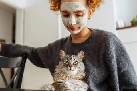 Gray fluffy tabby cat sits on arms teenage girl wearing cosmetic mask Stock Photos