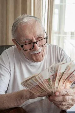 Gray-haired 88-year-old man with glasses happily counts his savings in rubles Stock Photos
