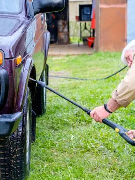 A gray-haired elderly man washes a car outdoors with a high-pressure apparatu Stock Photos