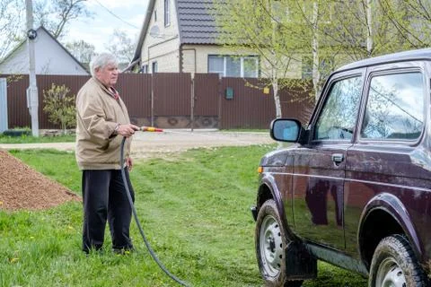 A gray-haired elderly man washes a car outdoors with a garden hose. Stock Photos