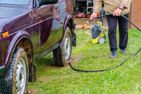 A gray-haired elderly man washes a car outdoors with a garden hose. Stock Photos